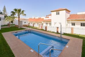 Una piscina con plataforma de baldosas, rodeada por un área cercada con palmeras y un edificio con techo de tejas.