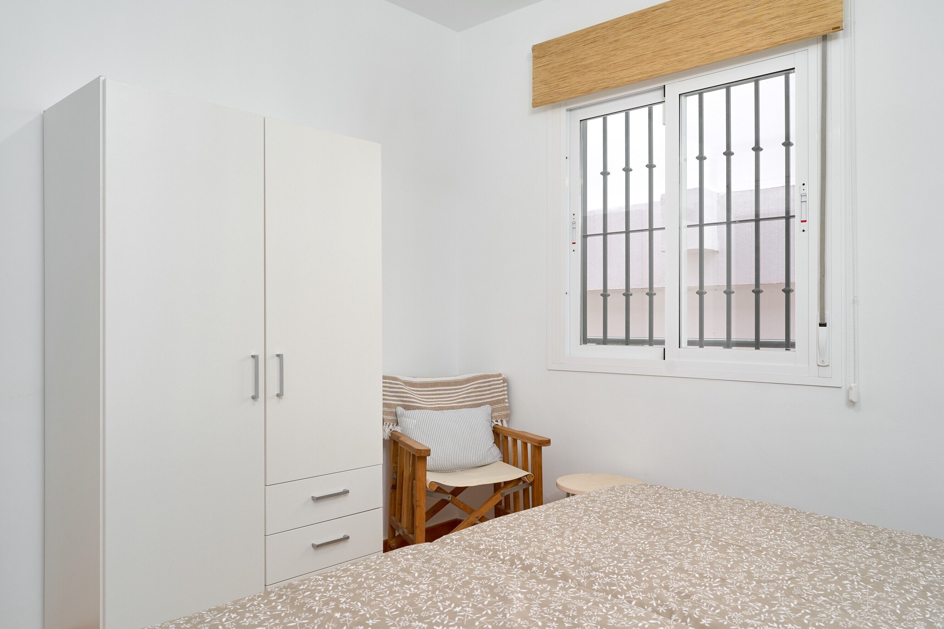 A bedroom with a bed, a wooden chair, a wardrobe, and a window with bars.