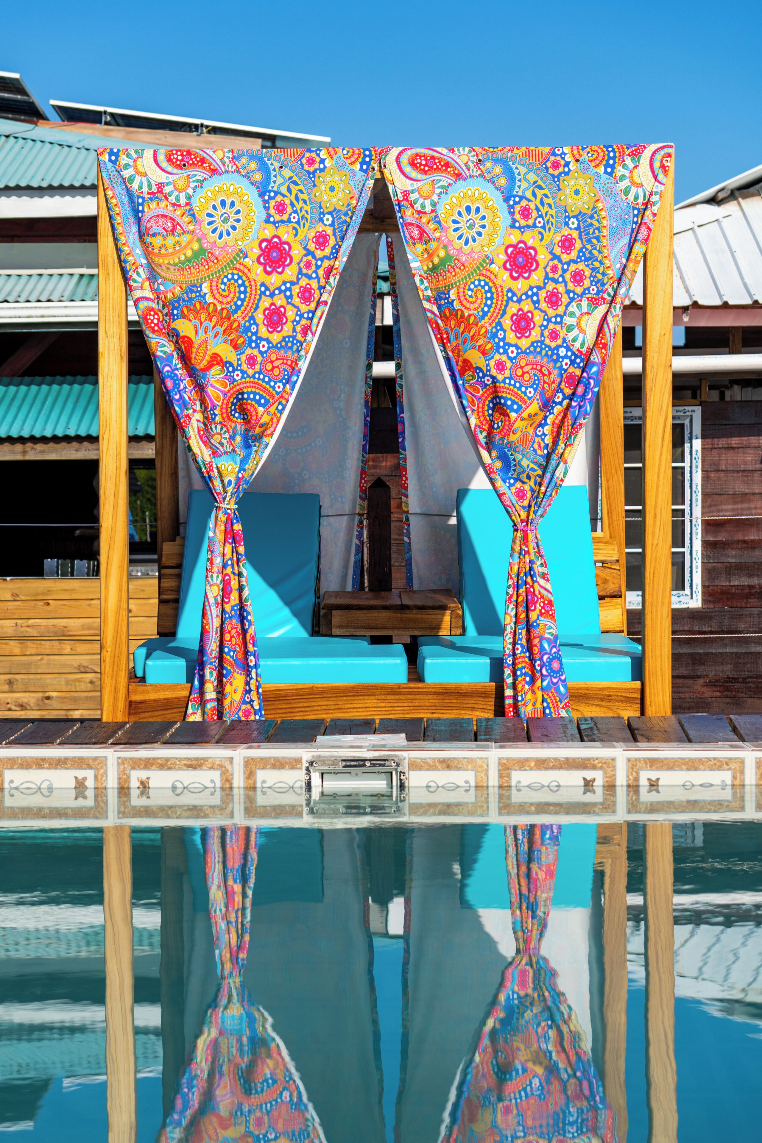 Un área de descanso junto a la piscina con cortinas coloridas y una estructura de madera.