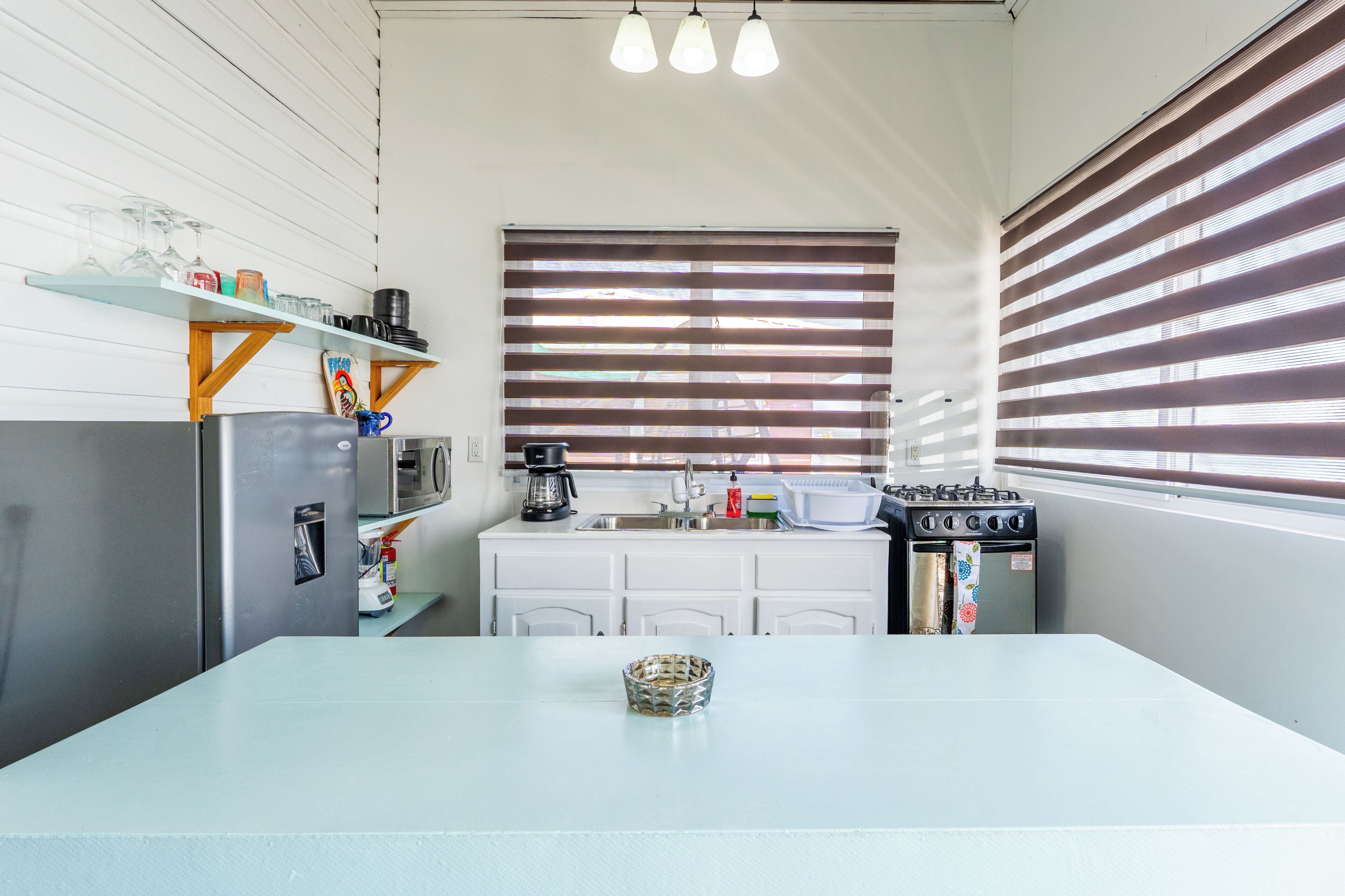 A compact kitchen with a refrigerator, stove, sink, and a window with blinds.