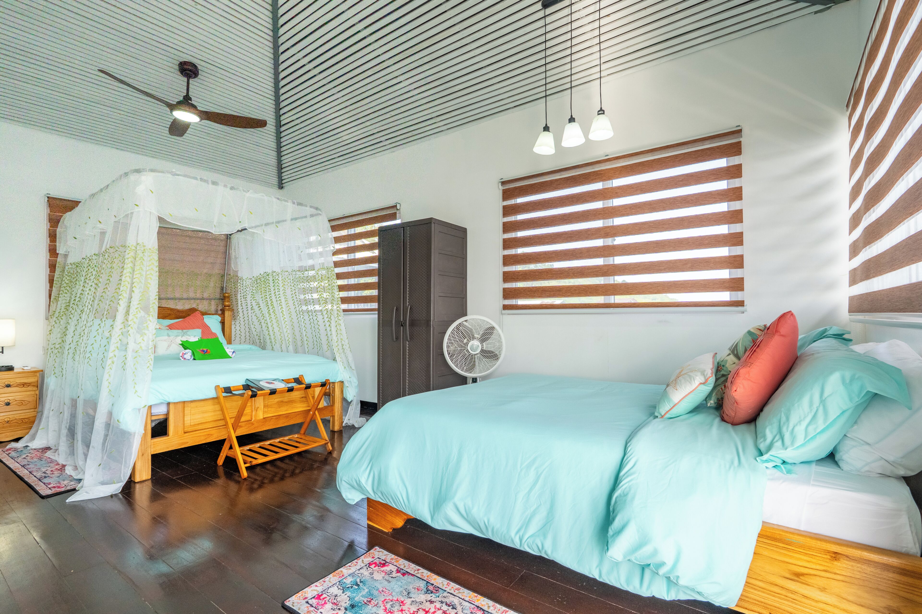 A bedroom with a bed, a ceiling fan, a standing fan, and a window with blinds.
