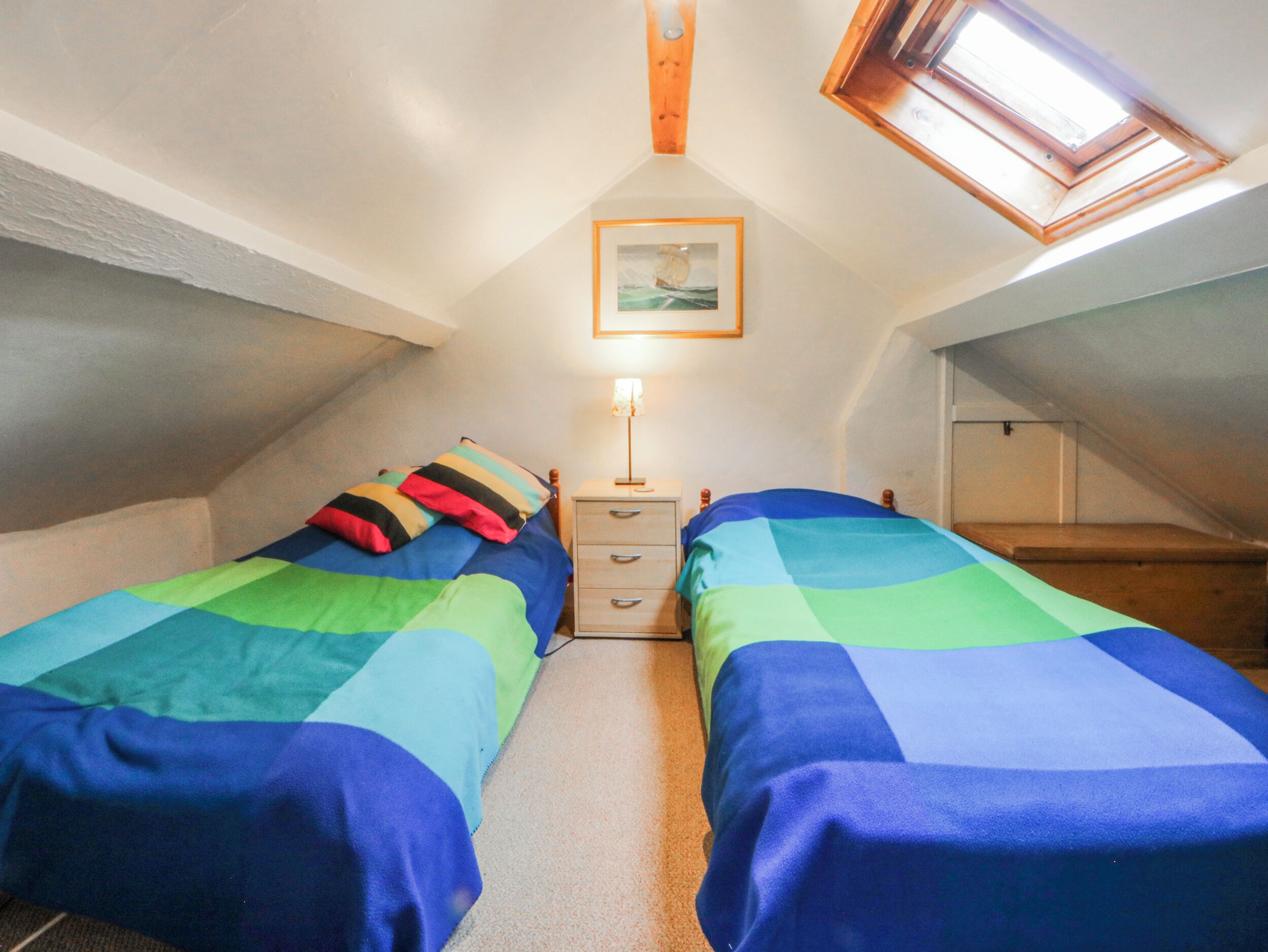Une petite chambre mansardée, chaleureuse et accueillante, avec deux lits, une table de chevet, une lampe et une fenêtre de toit.