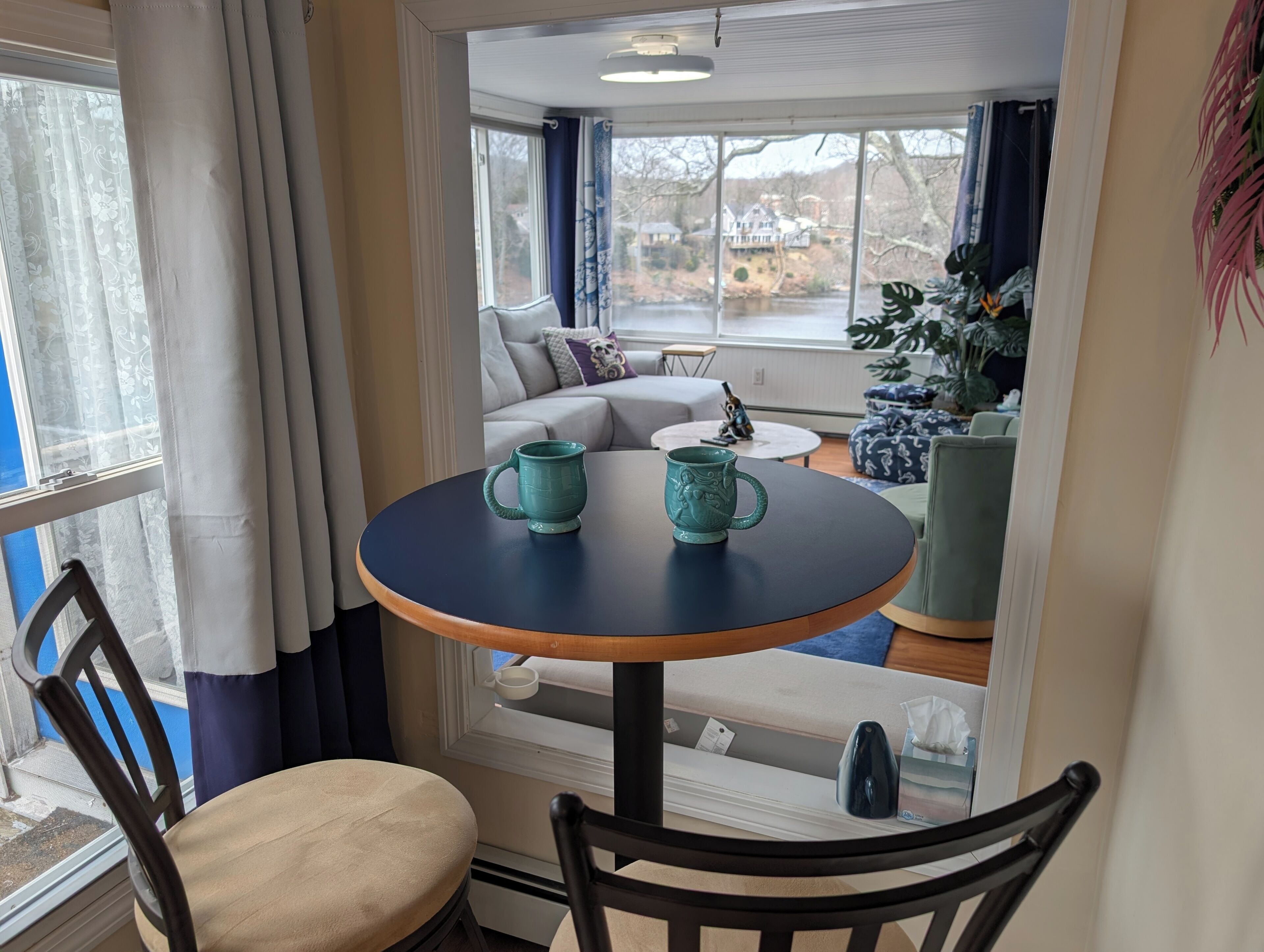 A dining area with a round table, two teal mugs, a chair, a sofa, and a view of a house and trees.