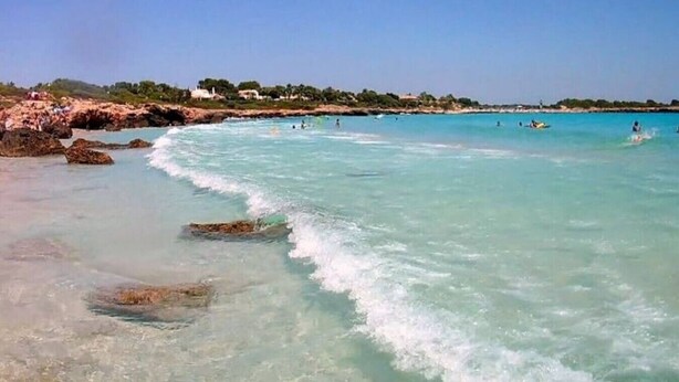 Een strand met helder turquoise water, een rotsachtige kustlijn en gebouwen in de verte.