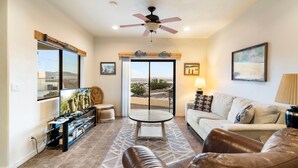 A living room with a television, leather sofa, a coffee table, and a ceiling fan. - Secluded Cottage with Fire-pit & Pool (Lake Havasu City)