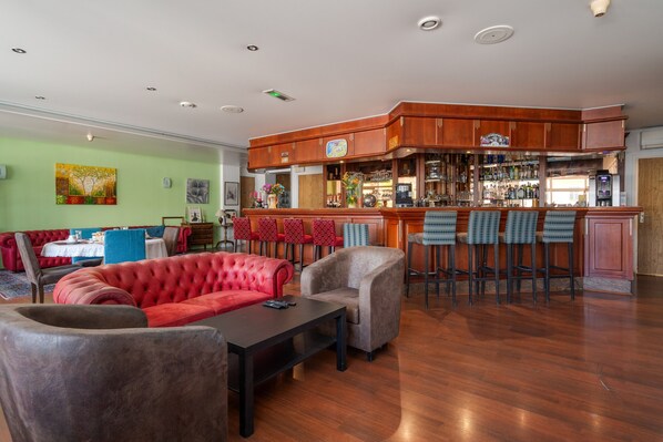 A bar area with wooden cabinets, a bar counter, and multiple stools. There is a red upholstered sofa and a table with chairs in the foreground.