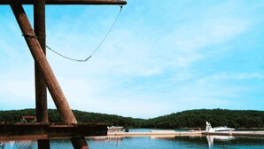 Eine Holzkonstruktion mit Kette, einem Steg und einem Boot auf ruhigem Gewässer, im Hintergrund ein bewaldeter Hügel.