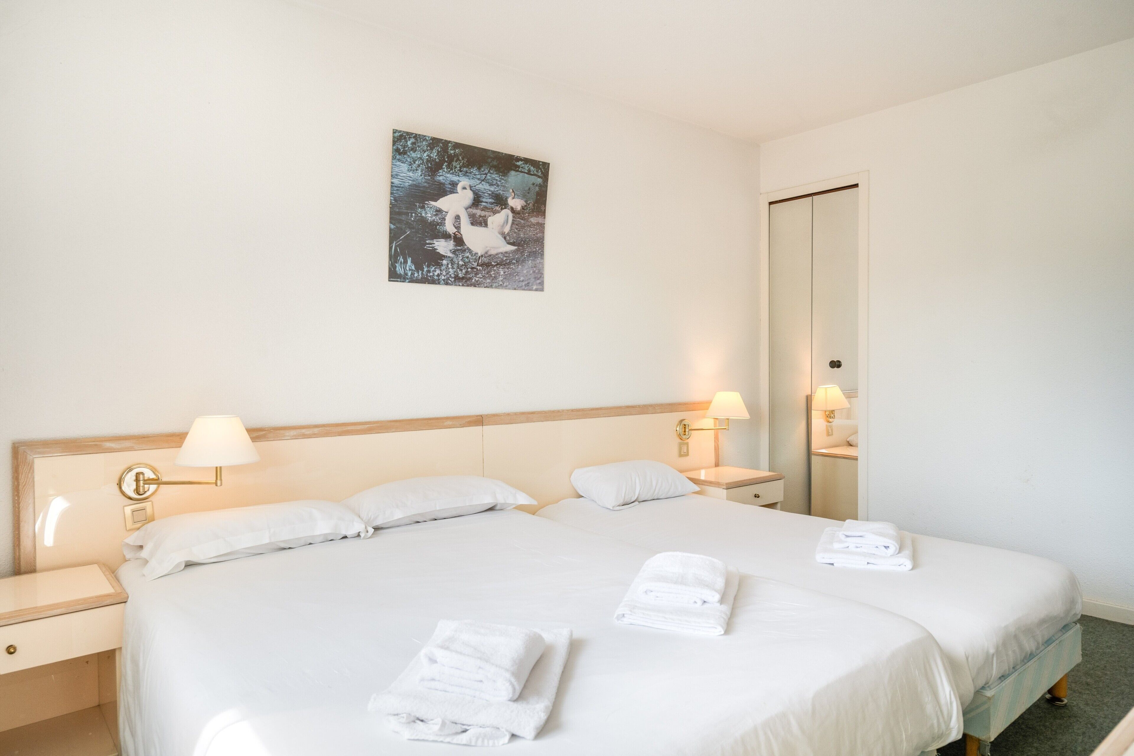 Een tweepersoonsbed met witte lakens, twee nachtkastjes met lampen, een kunstwerk aan de muur en een spiegel.
