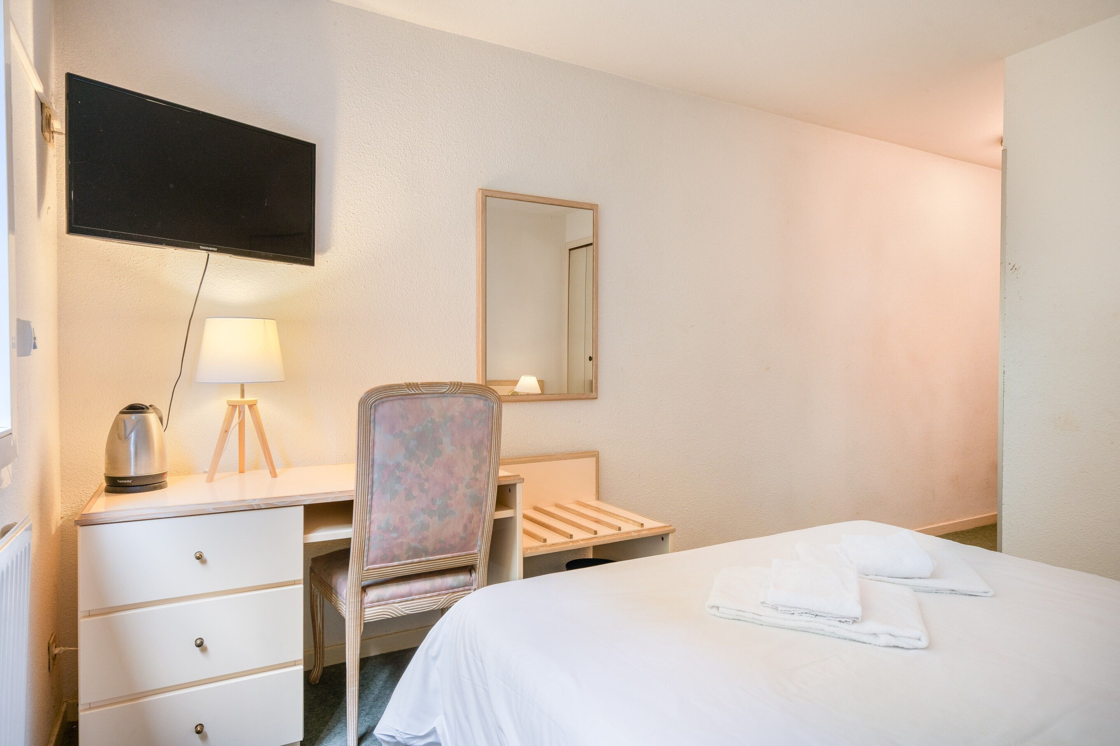 A hotel room with a bed, a desk, a chair, a lamp, a mirror, and a television.