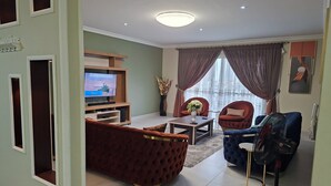 A living room with a TV, red and blue sofas, a table, and a standing fan. - Delightful 3-bedroom apartment in welcoming Accra neighborhood (Accra)