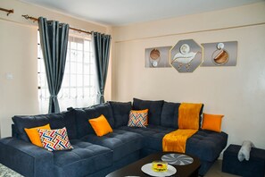 A living room with a dark blue sectional sofa, orange and patterned throw pillows, a coffee table, and wall art. - Cozy 1-bedroom apartment in Nairobi perfect for your stay (Nairobi)