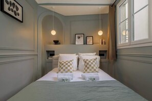 Uma cama bem arrumada com almofadas xadrez, uma mesinha de cabeceira com abajur e quadros nas paredes.
