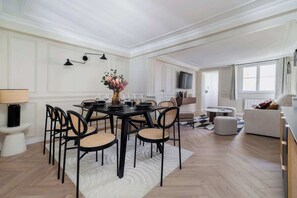 Ein Essbereich mit einem für vier Personen gedeckten Tisch, umgeben von Stühlen, einem Fernseher im Hintergrund und einem Sofa im angrenzenden Wohnbereich.