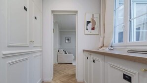 Un couloir avec des meubles blancs, un parquet en bois disposé en chevrons, une fenêtre avec des rideaux et une œuvre d’art encadrée accrochée au mur.