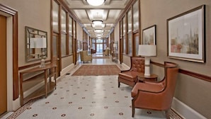 A hotel hallway with a patterned floor, leather armchairs, a desk with a lamp, framed artwork, and a large window at the end.