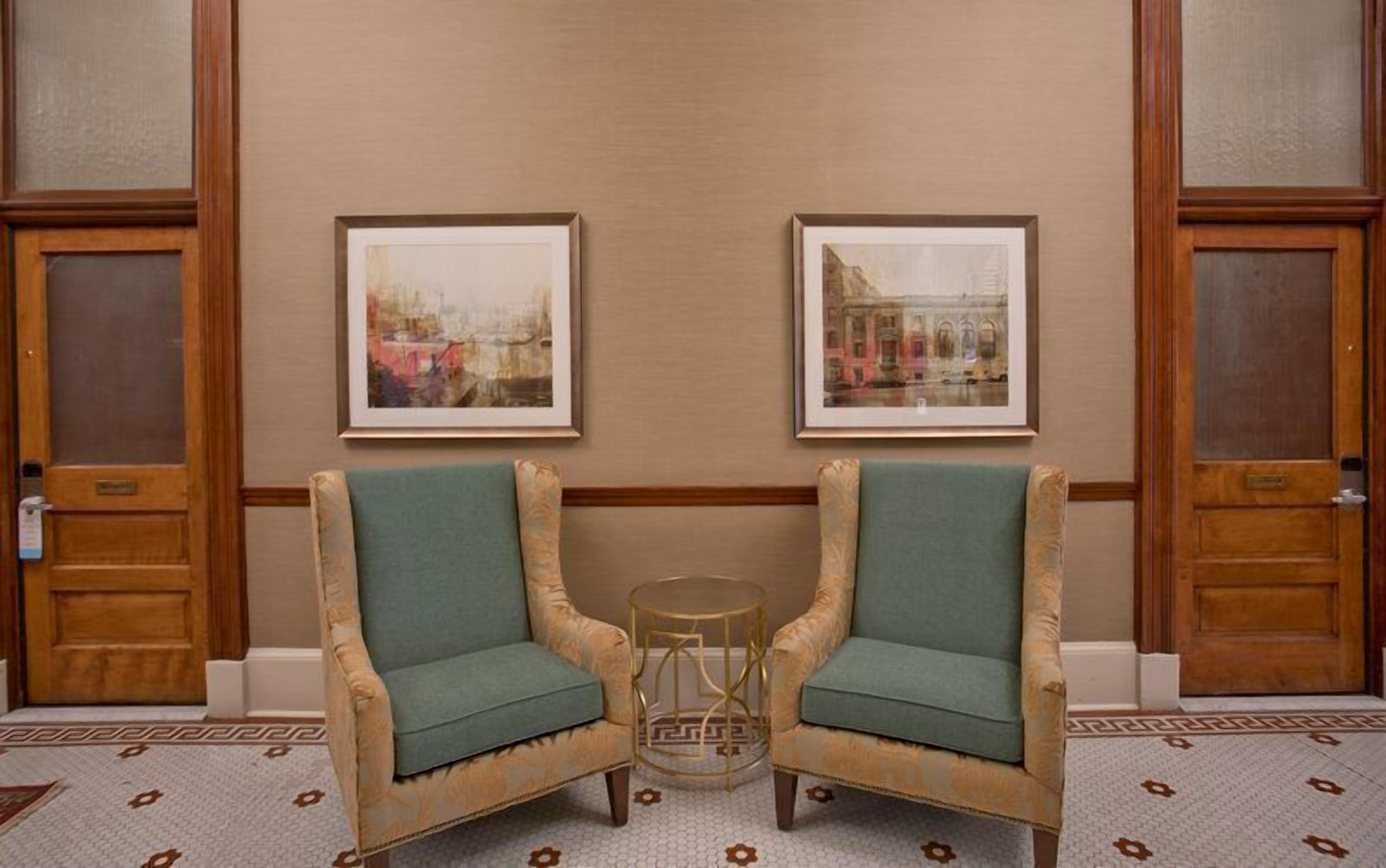 A hotel lobby with two armchairs, a small round table, and two framed pictures on the wall.