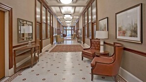 Ein Hotelgang mit einem gemusterten Boden, Ledersesseln, einem Schreibtisch mit Lampe, gerahmten Kunstwerken und einem großen Fenster am Ende.
