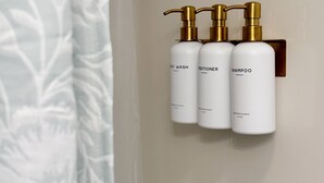 Trois flacons blancs avec des pompes dorées, portant les inscriptions « GEL DOUCHE », « APRÈS-SHAMPOING » et « SHAMPOO », sont fixés au mur.
