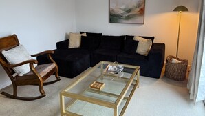 Un salon comprenant une table basse en verre, un fauteuil à bascule en bois, un canapé de couleur foncée, un tableau abstrait accroché au mur et une lampe sur pied.