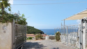 Une entrée fermée par un portail mène à une zone dallée de pierres, d’où l’on peut admirer la mer et les collines.