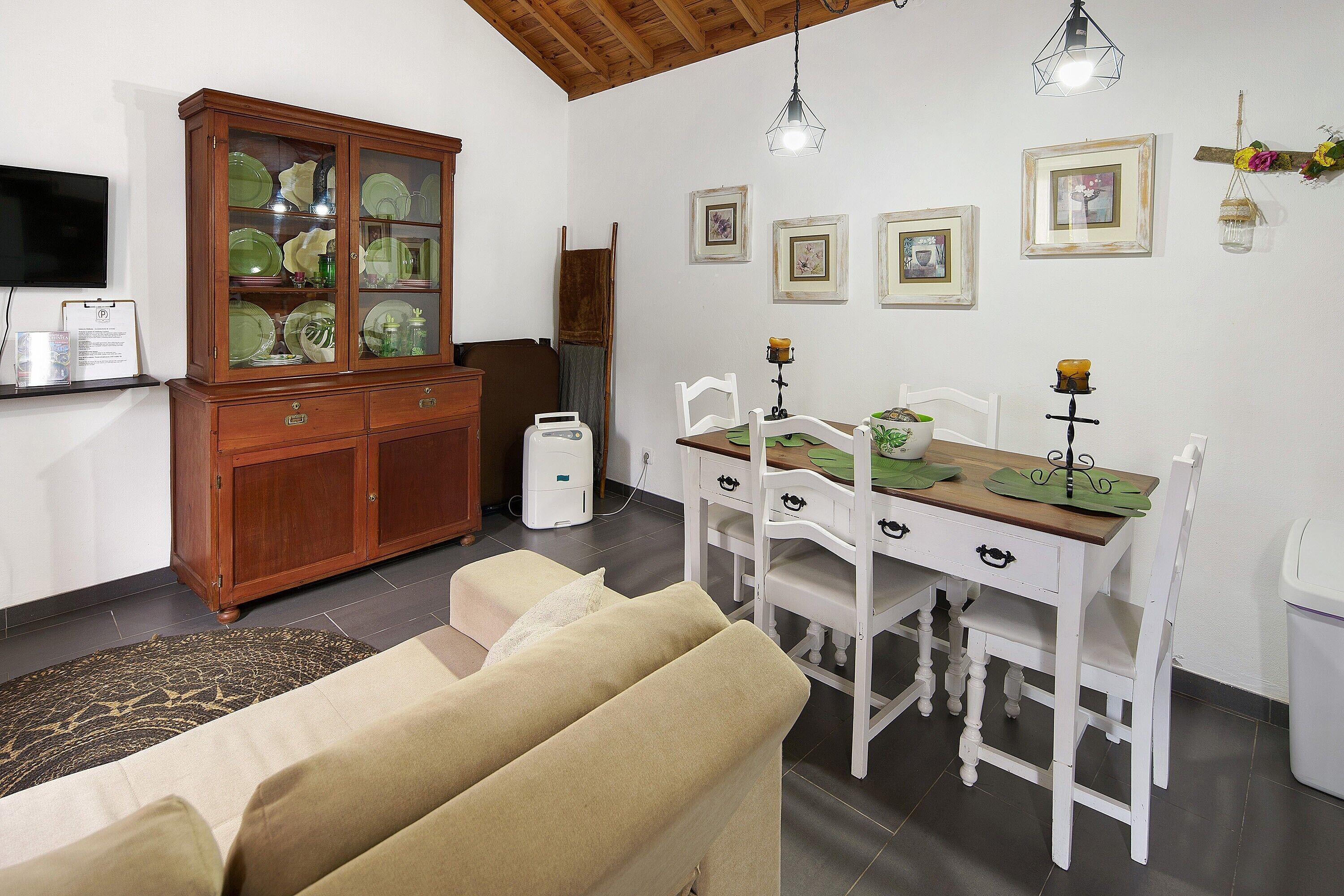 A dining area with a white table and chairs, a cabinet with glass doors, a television on the wall, and framed pictures on the wall.