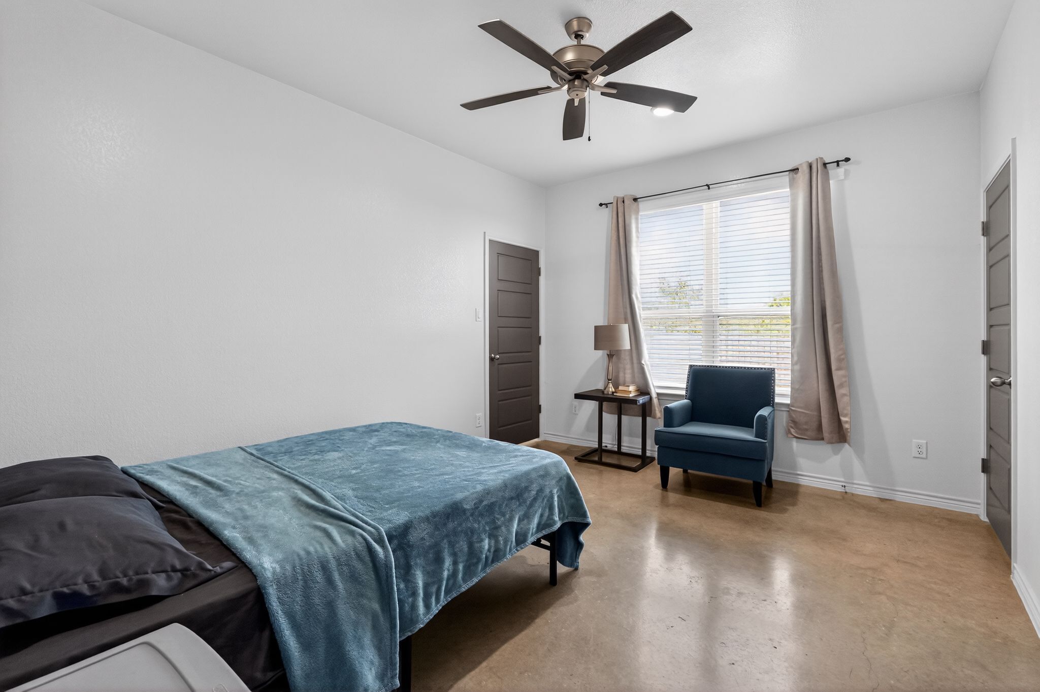 Un dormitorio con cama, una silla, ventilador de techo, ventana con cortinas y una puerta.