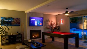 Een woonkamer met een open haard, een pooltafel, een televisietoestel en een plafondventilator.