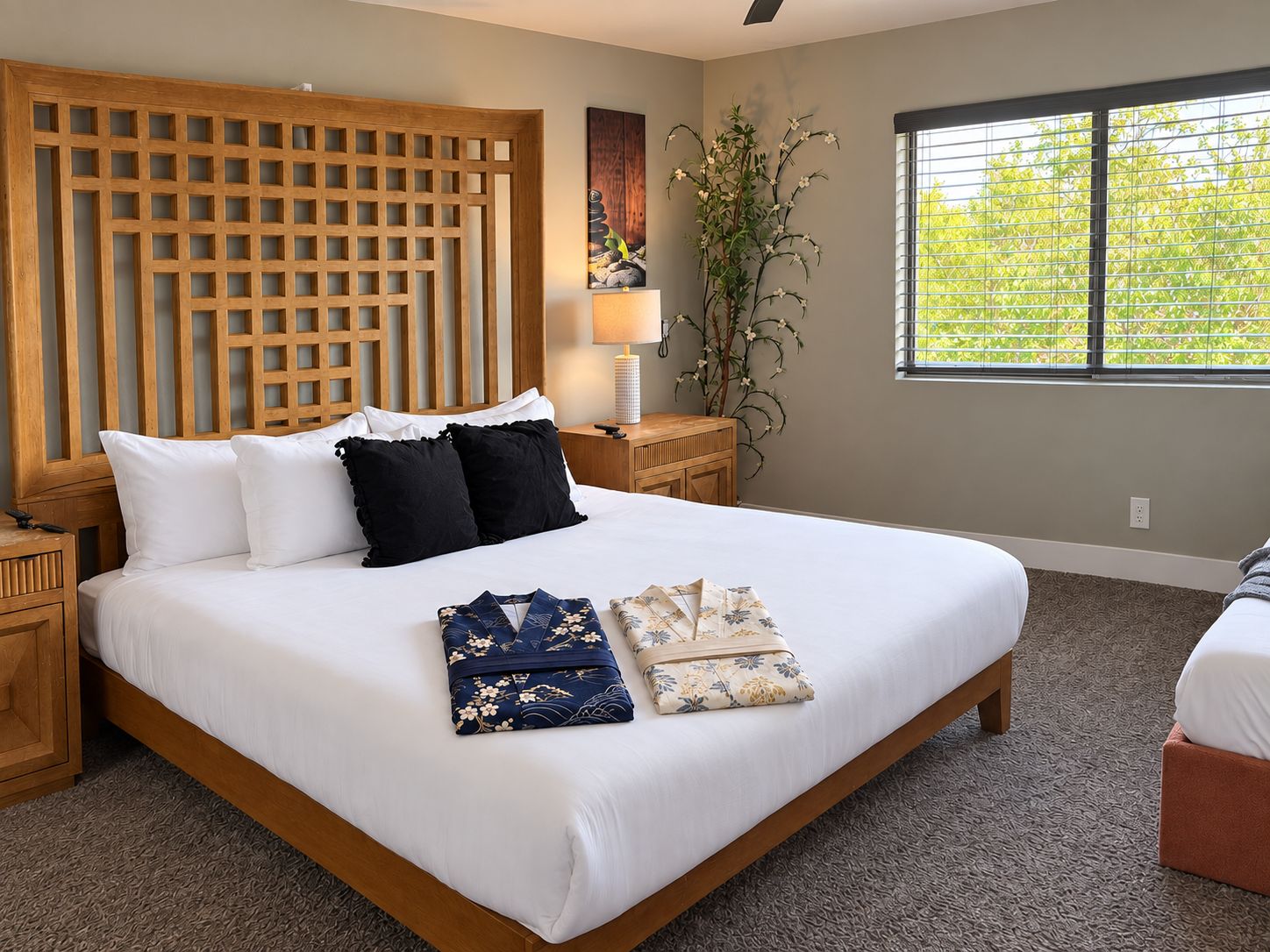A bedroom with a large bed, wooden headboard, bedside tables, a lamp, a potted plant, and a window with blinds.