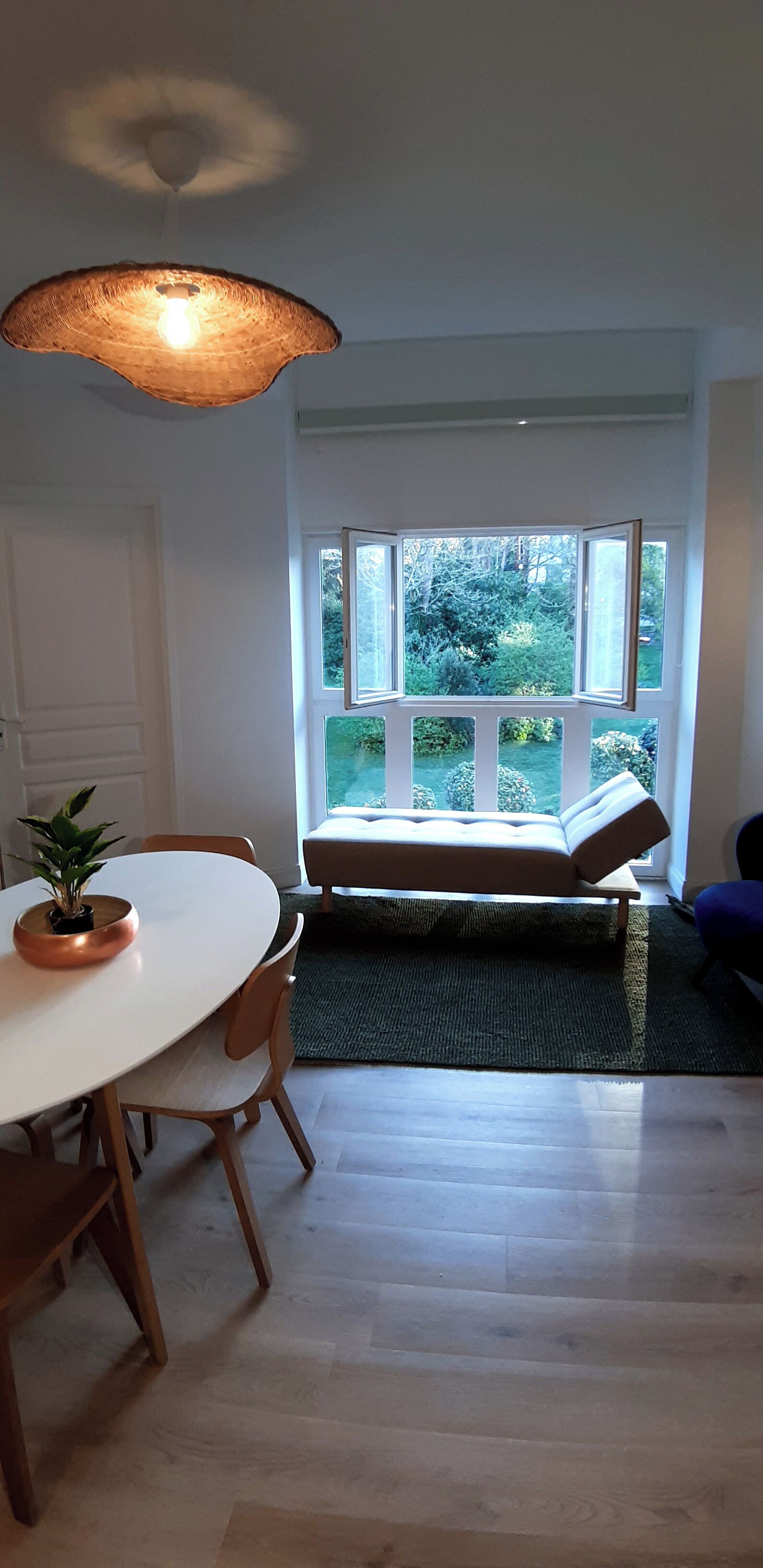 A room with a white table, chairs, a bench, a potted plant, and a view of greenery through a large window.