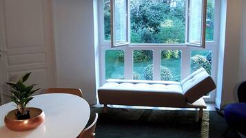 A room with a white table, chairs, a bench, a potted plant, and a view of greenery through a large window.