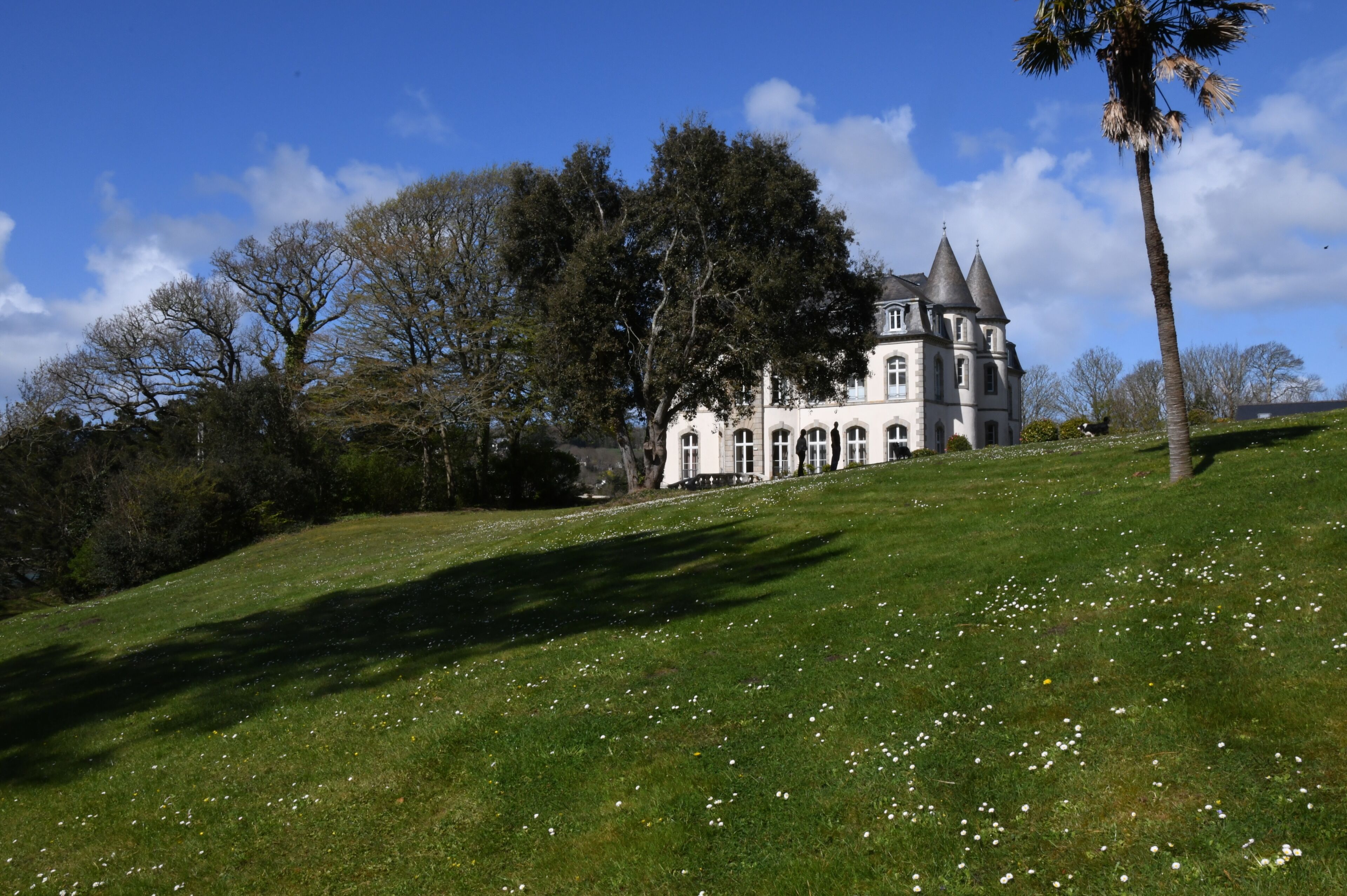 A large, historic building with a prominent tower, surrounded by lush greenery and a clear sky.