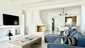 A modern living room with a TV, sofa, coffee table, fireplace, and kitchen in the background. - Villa Greystone & Sky Mykonos (Μύκονος)