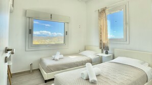 A hotel room with two beds, a large window with a city view, a ceiling fan, and a wall-mounted air conditioner. - Villa Greystone & Sky Mykonos (Μύκονος)