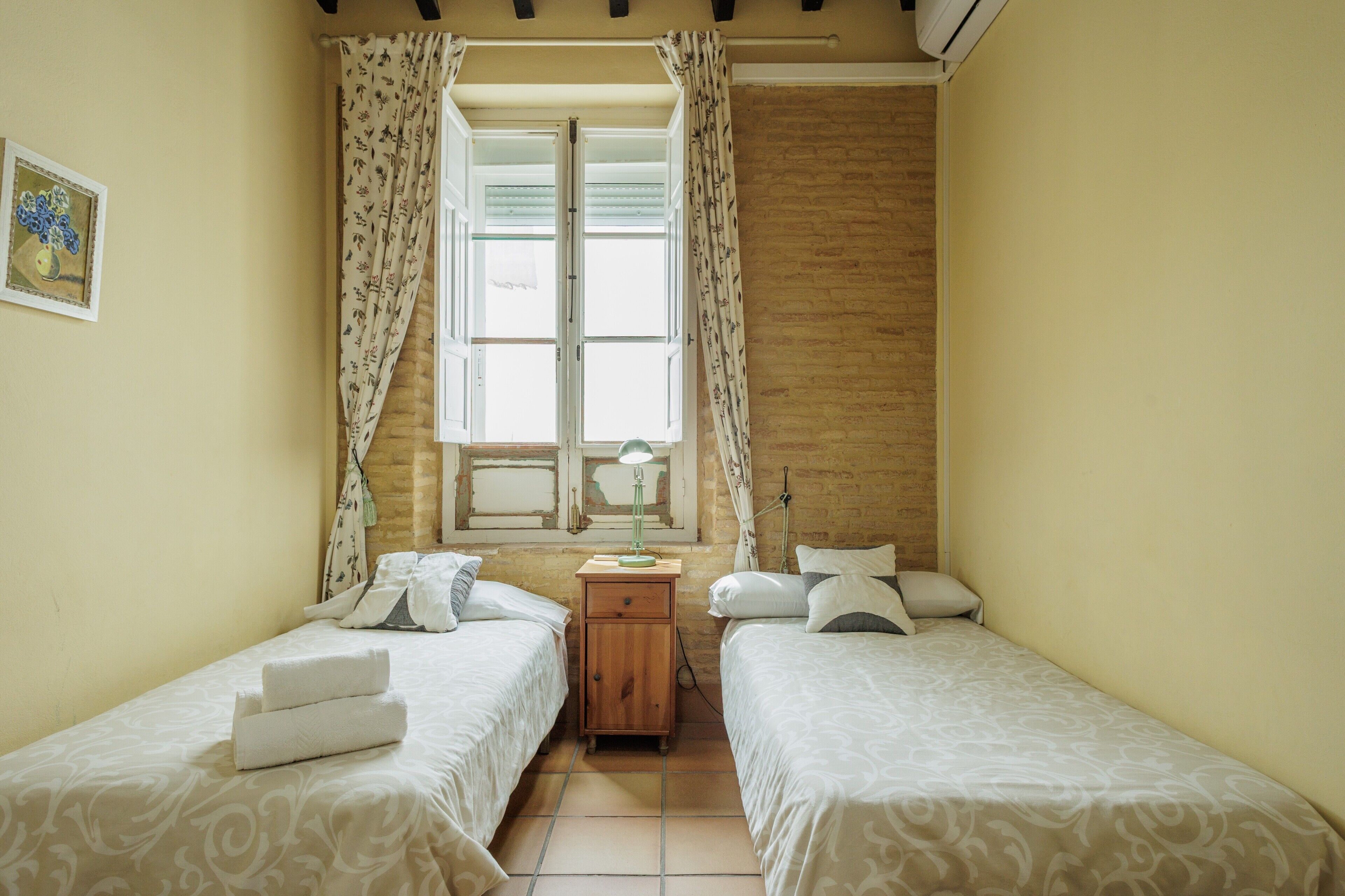 A room with two beds, a window with curtains, a bedside table with a lamp, and a wall-mounted air conditioner.
