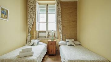 A room with two beds, a window with curtains, a bedside table with a lamp, and a wall-mounted air conditioner.