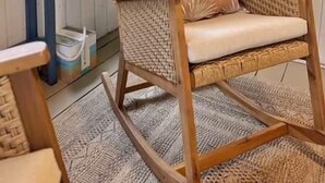 A wicker rocking chair with a cushion, a small table with a patterned lamp, and a large plant in a room with wooden walls and a window. - Mon Sherri - The Cottage (Loxley)