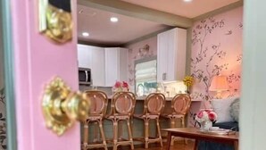 A pink door with a brass handle, a dining area with a wooden table and chairs, a kitchen in the background, and a patterned wall. - Mon Sherri - The Cottage (Loxley)