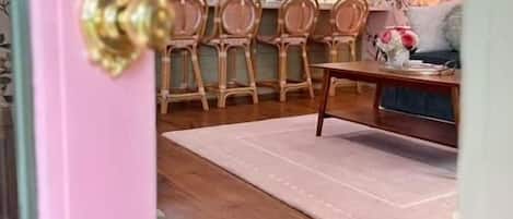 A pink door with a brass handle, a dining area with a wooden table and chairs, a kitchen in the background, and a patterned wall.