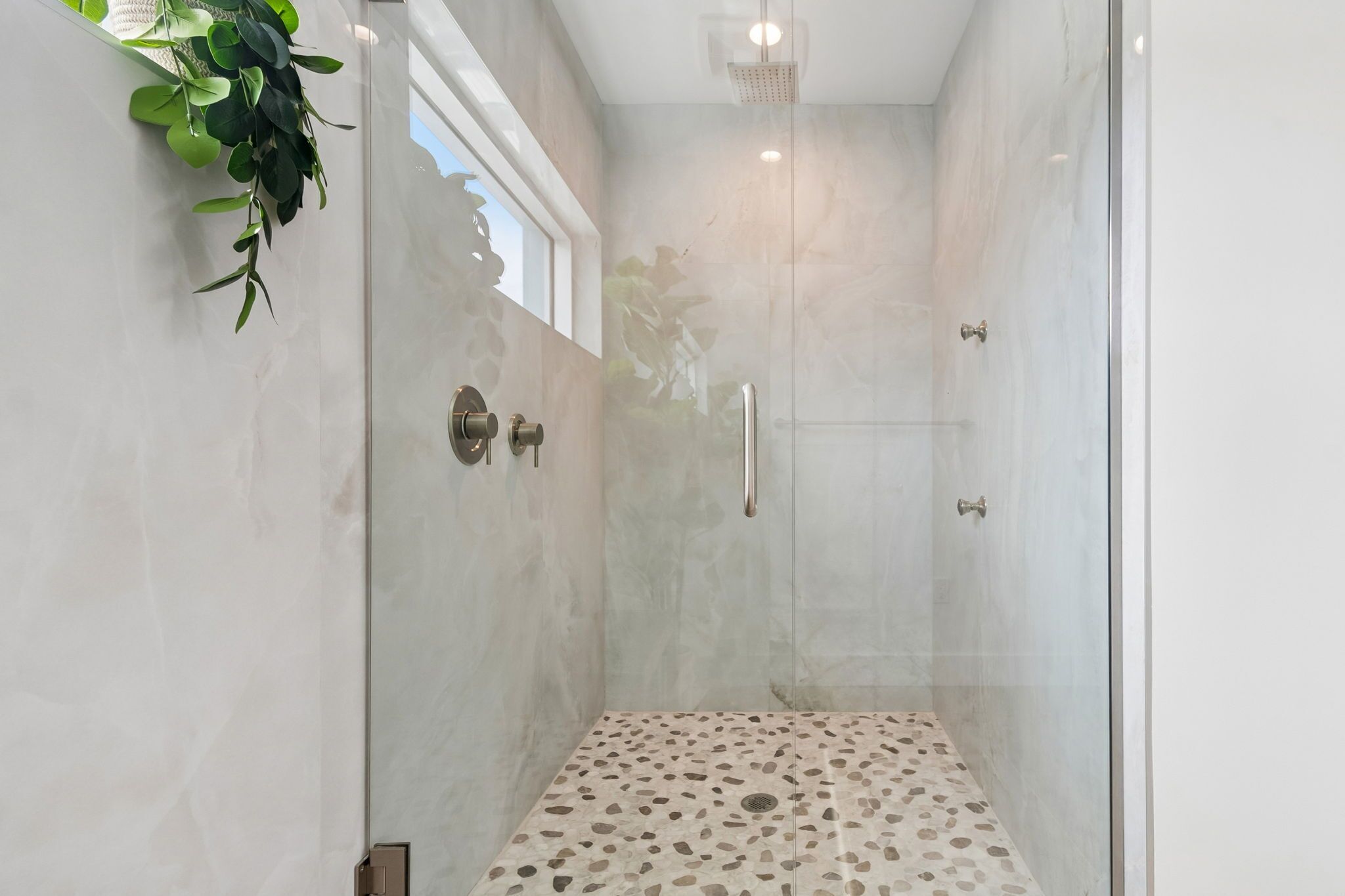 A glass-enclosed shower with a pebble-patterned floor, a window with a view, and a plant on the wall.