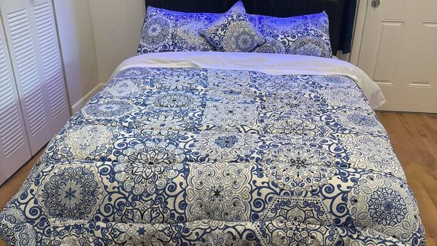Un lit avec une couvre-lit à motifs, une tête de lit dotée d’un éclairage bleu et une fenêtre avec des stores.
