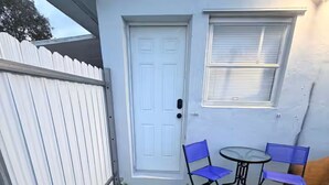 Une petite terrasse avec une porte blanche, deux chaises bleues et une petite table.