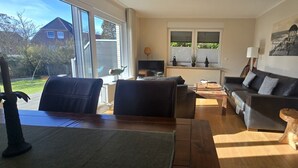 A living room with a sofa, coffee table, and a dining table. There is a window with a view of houses and trees.