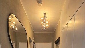 Un couloir étroit au plancher en bois, un miroir, une coiffeuse ornée d’un vase et une porte au fond.