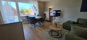 A modern living room with a sofa, a dining table with chairs, a TV on a stand, and a view through large windows. - Vacation apartment Ida (Borkum)