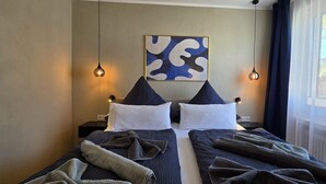 Un lit double avec une tête de lit, deux lampes de chevet, un tableau abstrait accroché au mur et une fenêtre avec des rideaux.