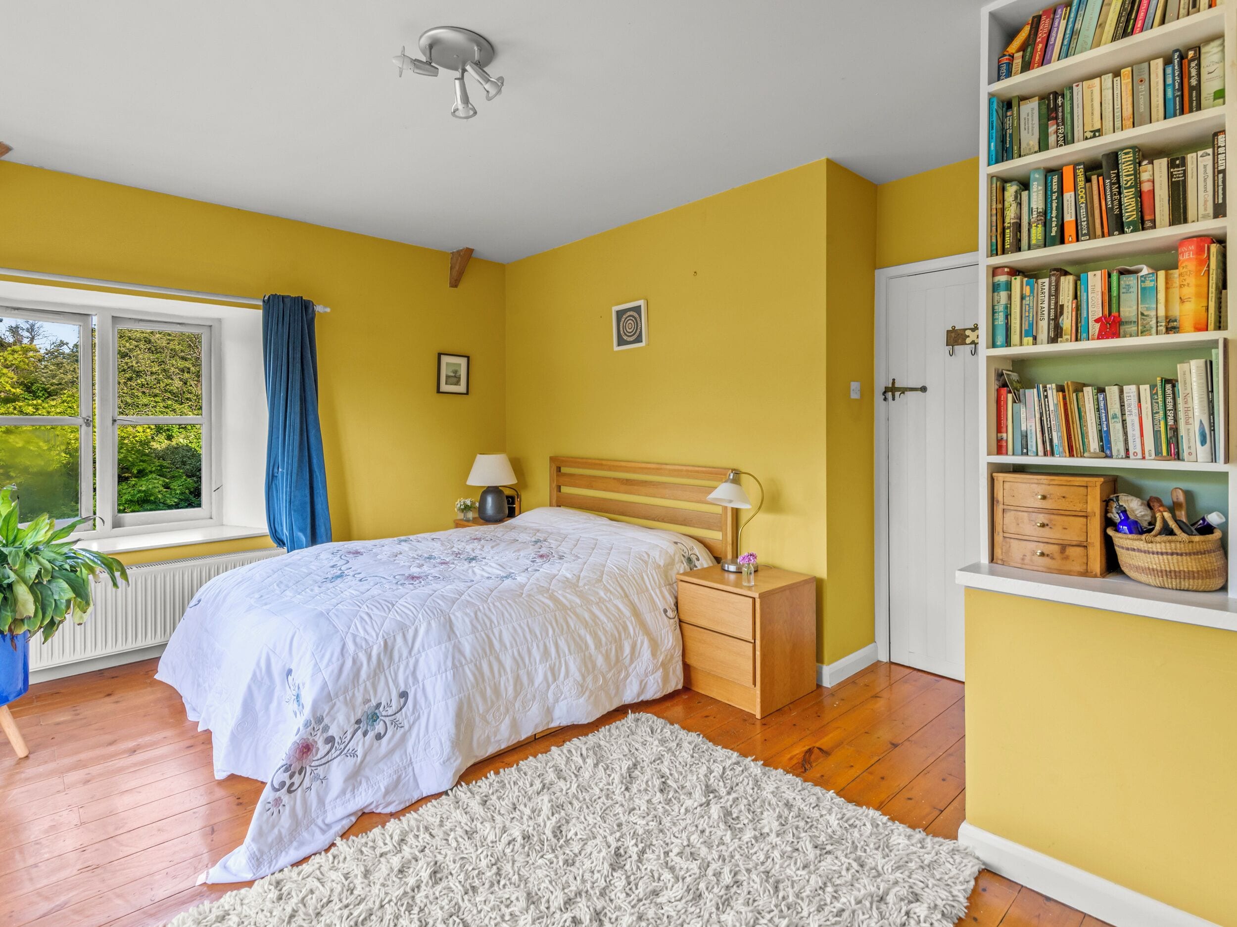 A bedroom with a bed, bedside tables, a bookshelf, a window with curtains, and a radiator.