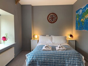 Ein Schlafzimmer mit einem Bett, zwei Nachttischen mit Lampen, einer Wanduhr, einer Karte und einem Fenster mit Blumen.
