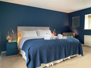 Een keurig opgemaakt bed met witte lakens, een blauw hoofdbord en een nachtkastje met een lamp en een bloemenvaas.