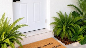 A white door with a number '1' sign above it, a doormat with a welcome message, and two potted ferns on either side of the door. - Casa Tropica Escape | 2BR Near Beach & Airport (Dania Beach)