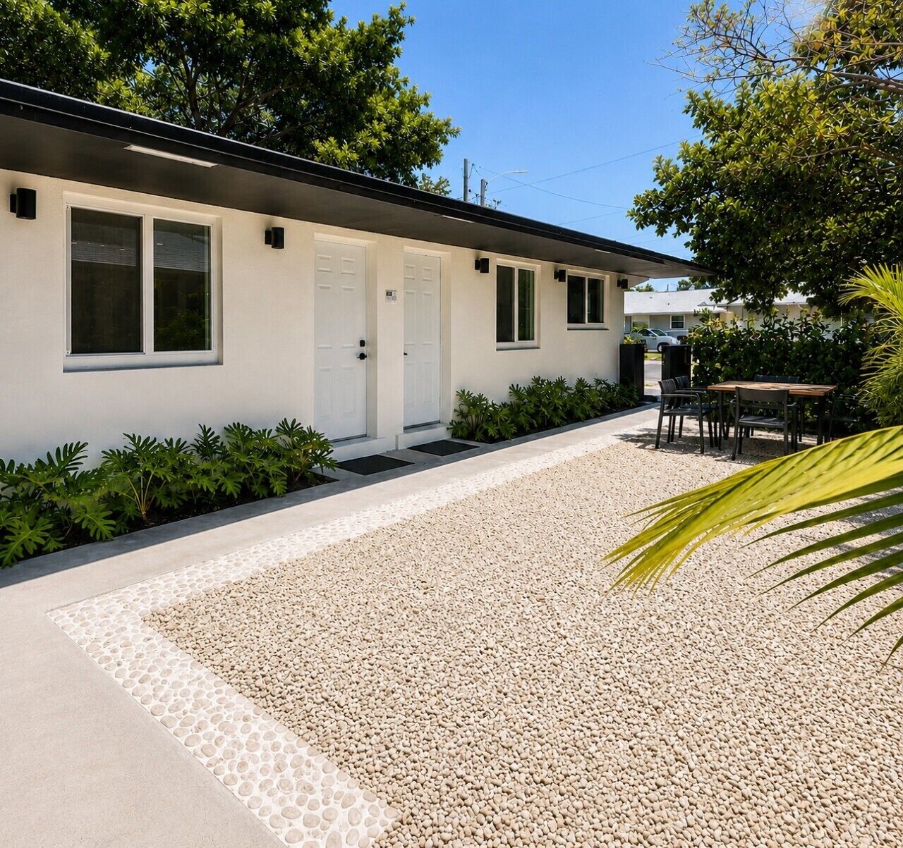 A modern building with a gravel driveway, a small patio with outdoor seating, and a row of potted plants along the side.